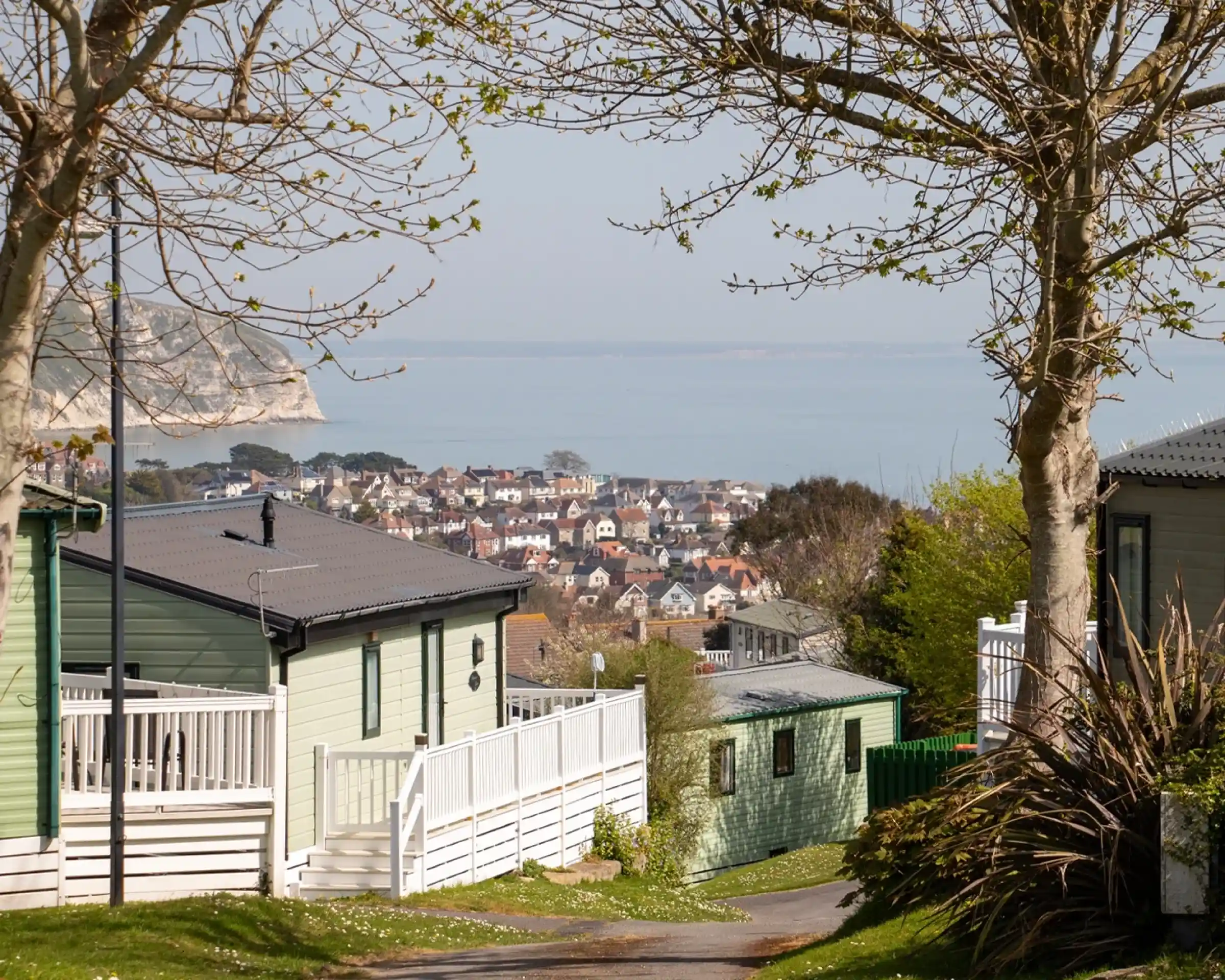 A scenic view of a coastal town from a hillside. The foreground features green and white residential buildings, with trees lining the pathway. In the background, the town's homes are visible along the water's edge, surrounded by calm sea and distant cliffs.