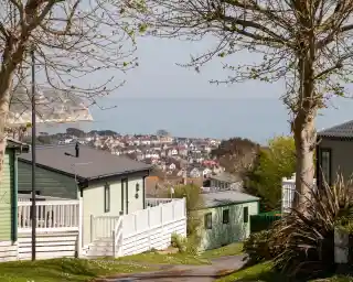 A scenic view of a coastal town from a hillside. The foreground features green and white residential buildings, with trees lining the pathway. In the background, the town's homes are visible along the water's edge, surrounded by calm sea and distant cliffs.