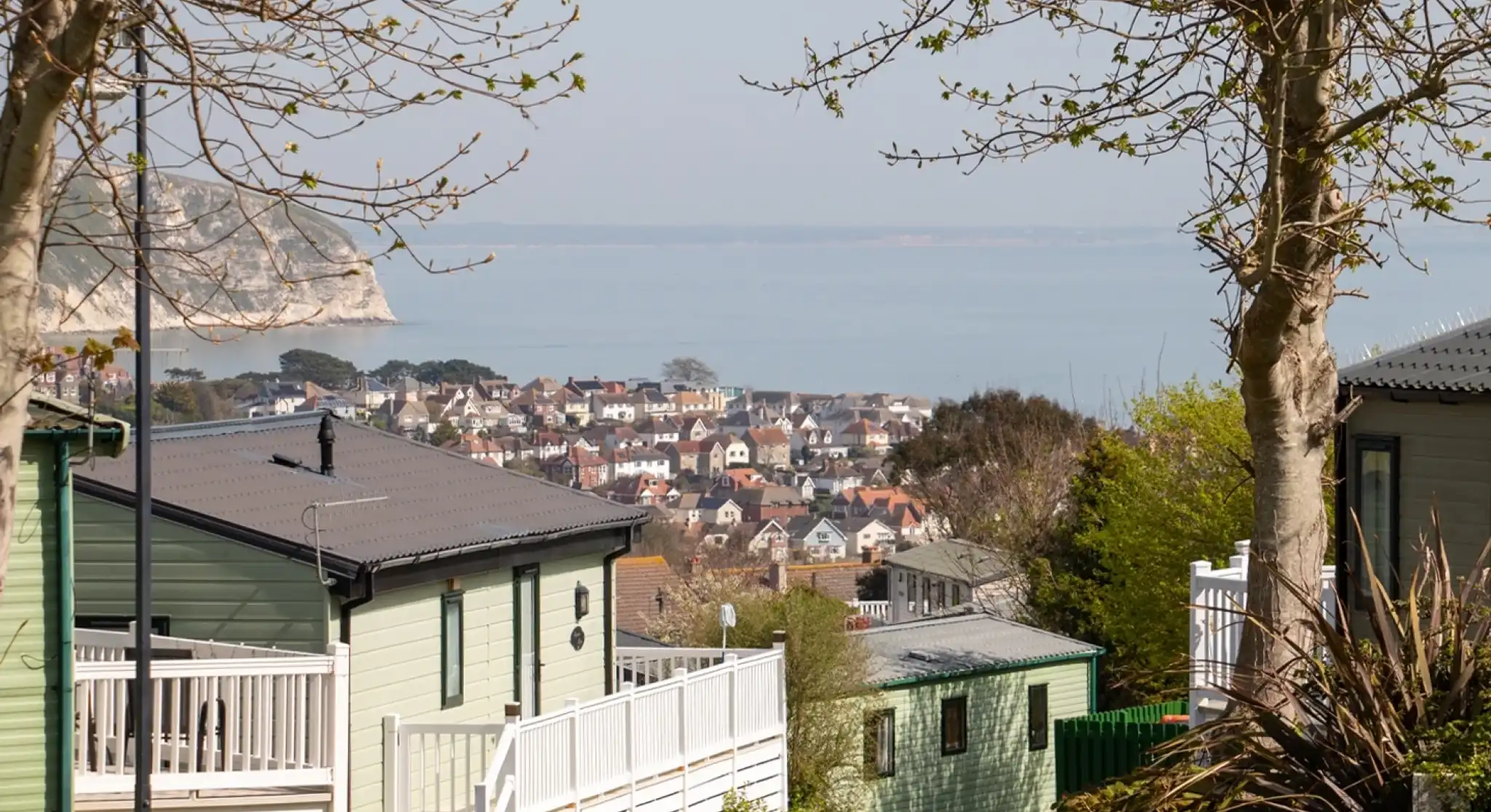 A scenic view of a coastal town from a hillside. The foreground features green and white residential buildings, with trees lining the pathway. In the background, the town's homes are visible along the water's edge, surrounded by calm sea and distant cliffs.