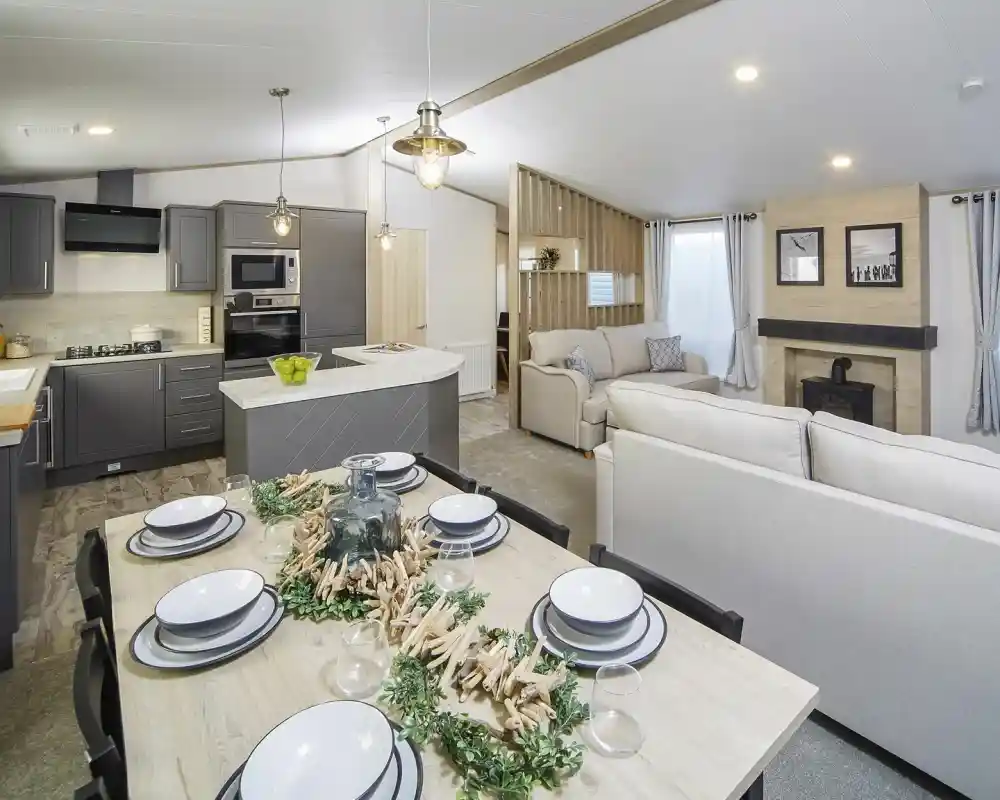 Modern open-plan kitchen, dining, lounge area in the Atlas Lilac Lodge with grey cabinetry, light wood dining table and chairs and neutral sofas.