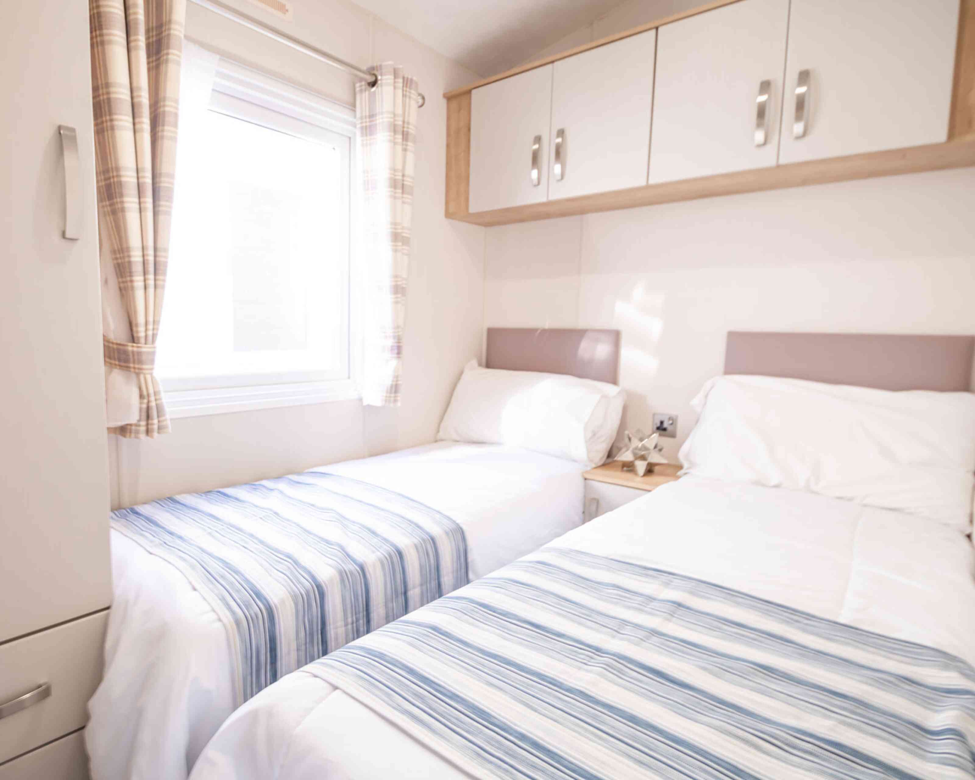 A bedroom with twin beds, overhead storage and wardrobe, together with a window.