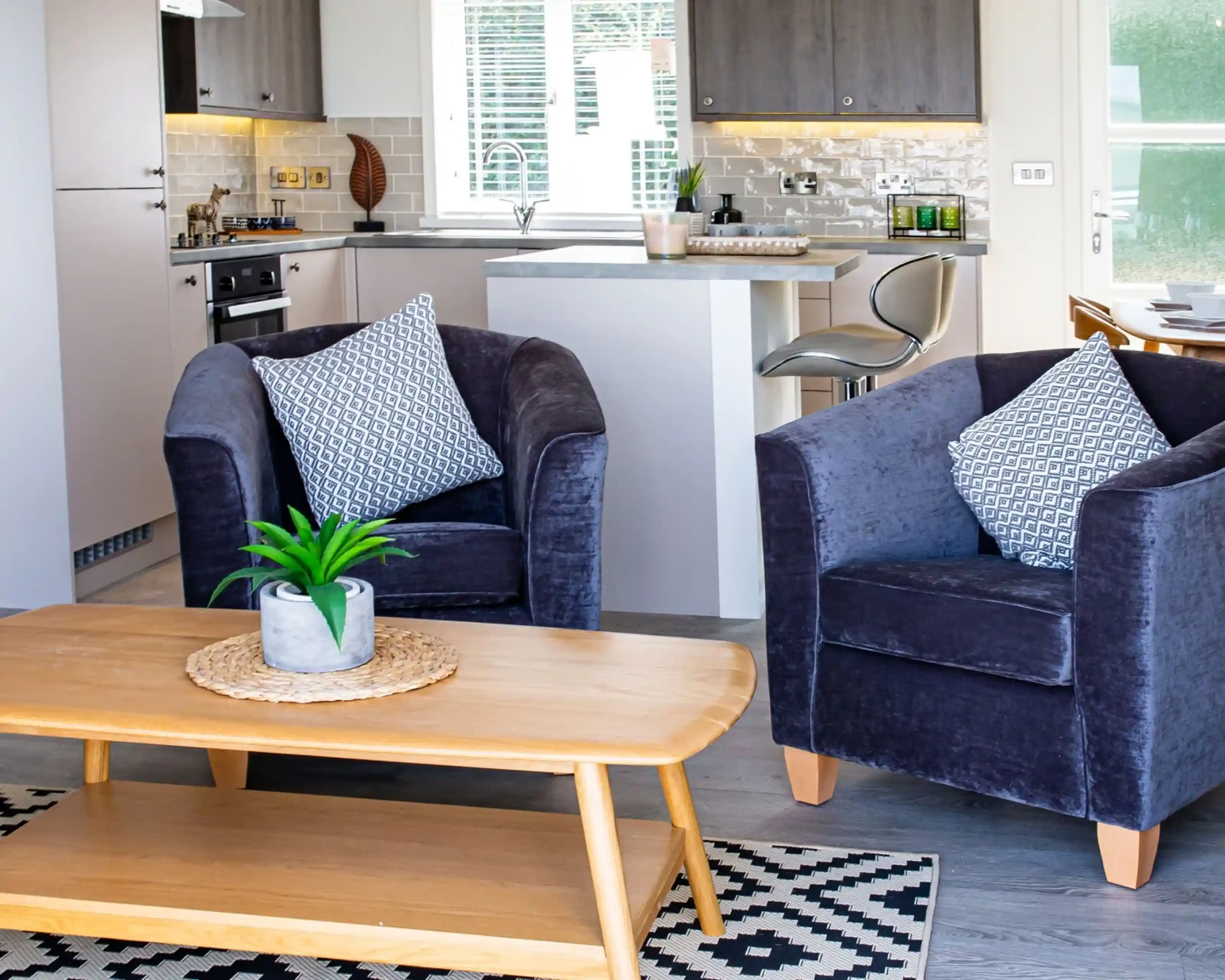 Cosy purple velvet tub chairs surrounding a light wooden coffee table with kitchen in the background