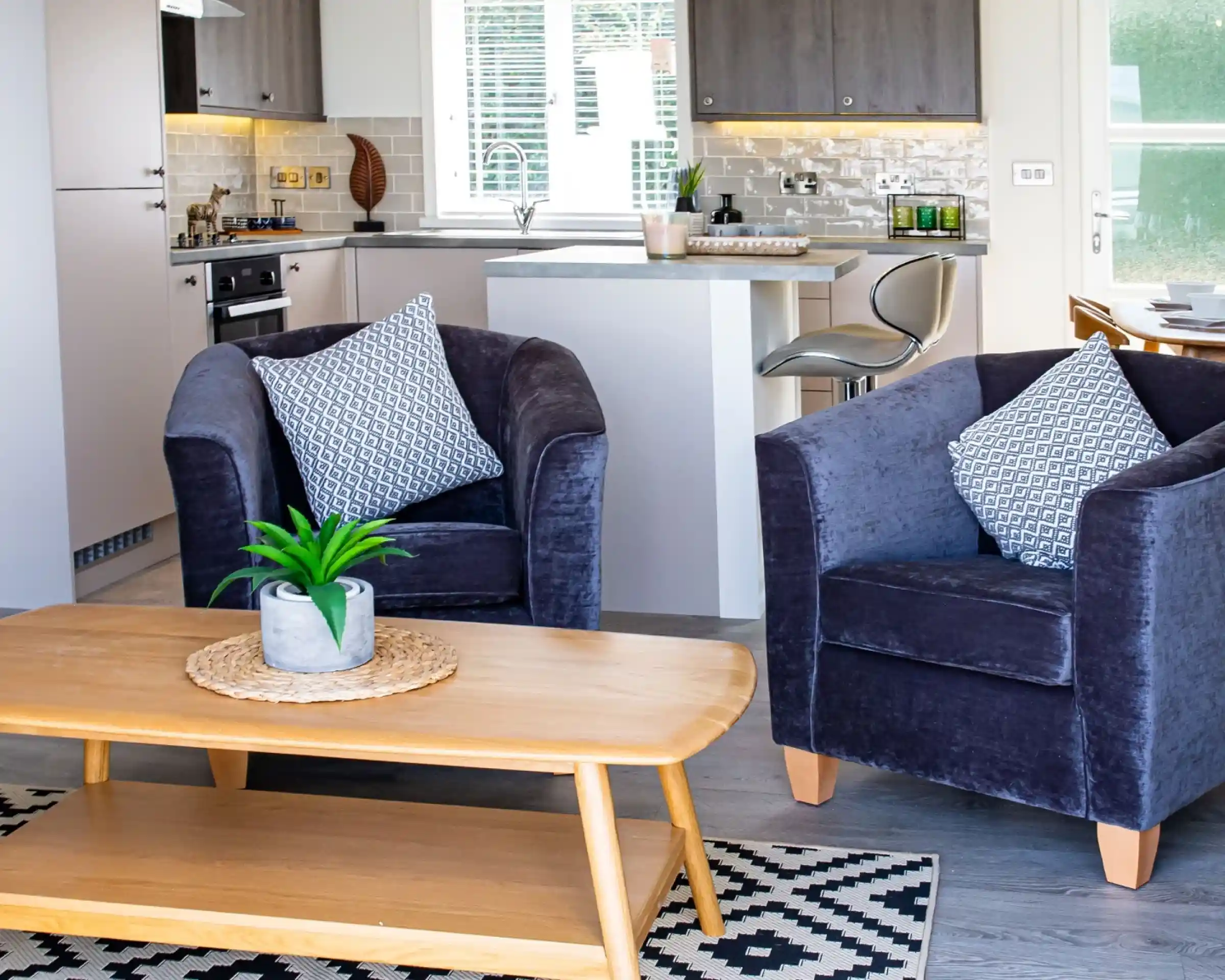 Cosy purple velvet tub chairs surrounding a light wooden coffee table with kitchen in the background