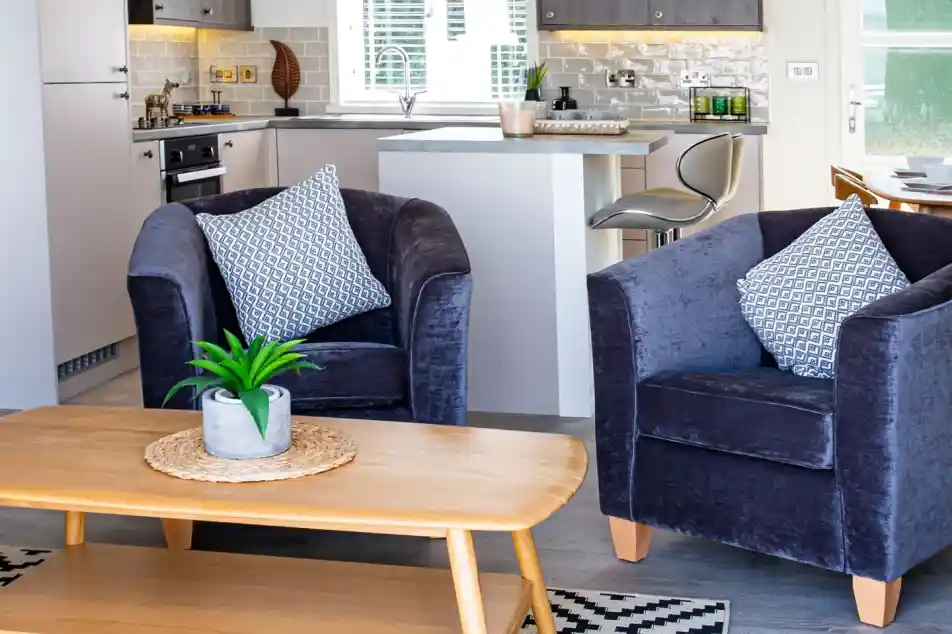 Cosy purple velvet tub chairs surrounding a light wooden coffee table with kitchen in the background