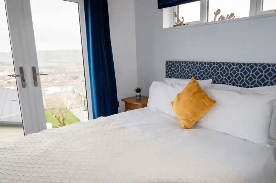 Double bed with white sheets and plush headboard in bedroom with patio doors and views over Swanage Coastal Park
