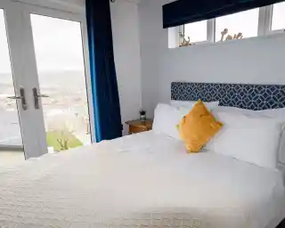 Double bed with white sheets and plush headboard in bedroom with patio doors and views over Swanage Coastal Park