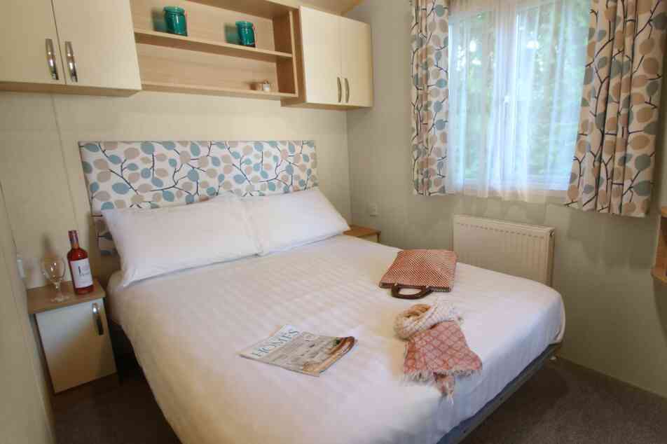 A cozy bedroom featuring a large bed with a patterned headboard. Light beige walls and curtains complement the room, which includes a bedside table with a wine bottle and glass. A folded blanket and a magazine are placed on the side of the bed. Natural light enters from a nearby window.