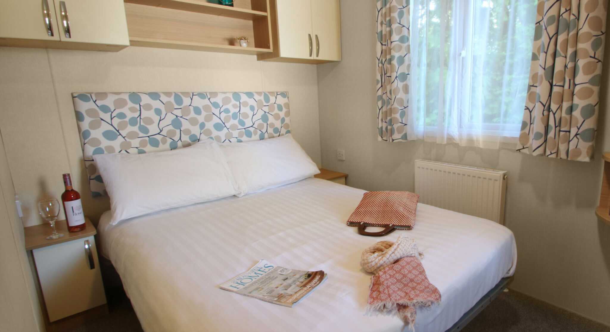 A cozy bedroom featuring a large bed with a patterned headboard. Light beige walls and curtains complement the room, which includes a bedside table with a wine bottle and glass. A folded blanket and a magazine are placed on the side of the bed. Natural light enters from a nearby window.