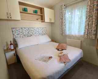 A cozy bedroom featuring a large bed with a patterned headboard. Light beige walls and curtains complement the room, which includes a bedside table with a wine bottle and glass. A folded blanket and a magazine are placed on the side of the bed. Natural light enters from a nearby window.