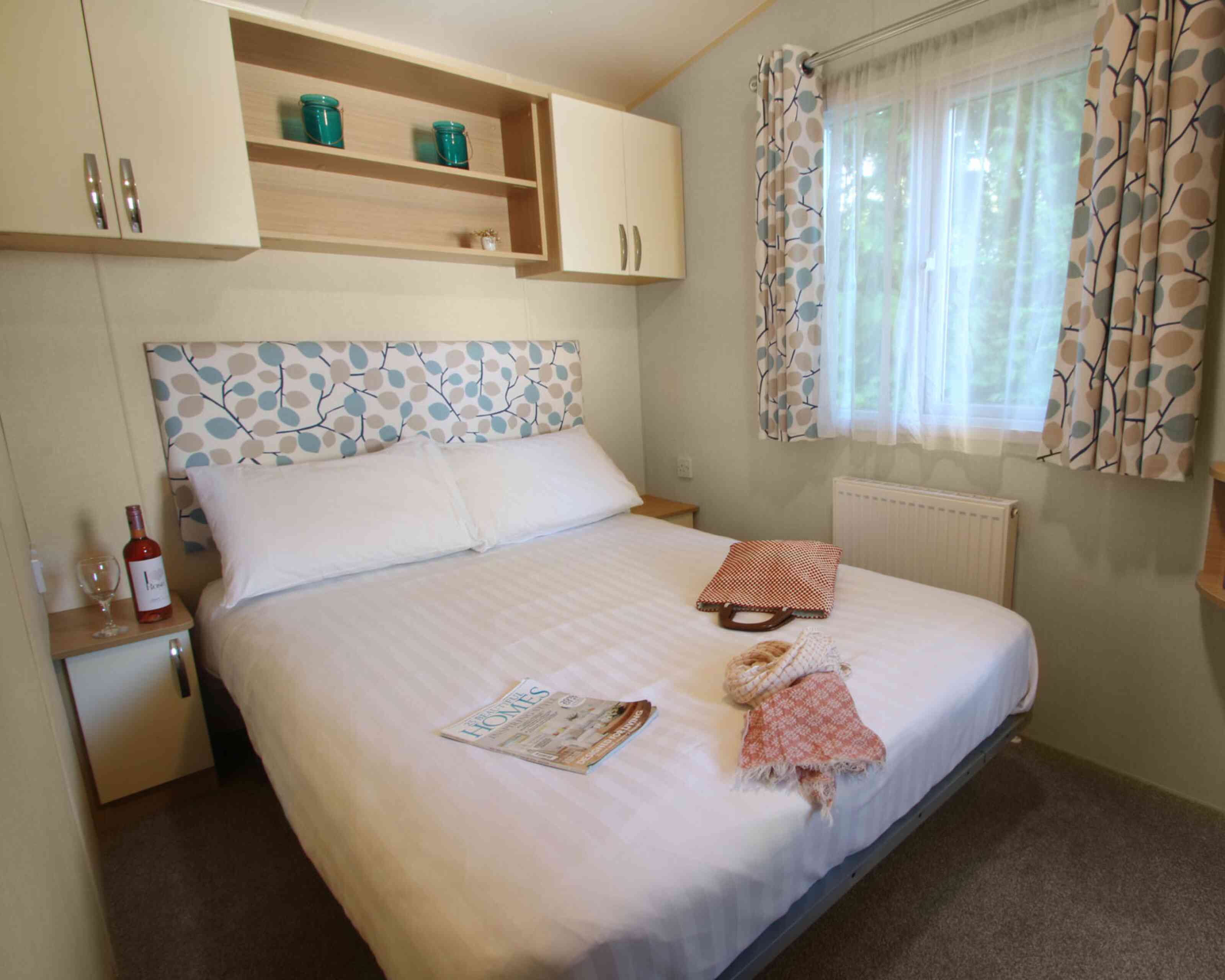 A cozy bedroom featuring a large bed with a patterned headboard. Light beige walls and curtains complement the room, which includes a bedside table with a wine bottle and glass. A folded blanket and a magazine are placed on the side of the bed. Natural light enters from a nearby window.