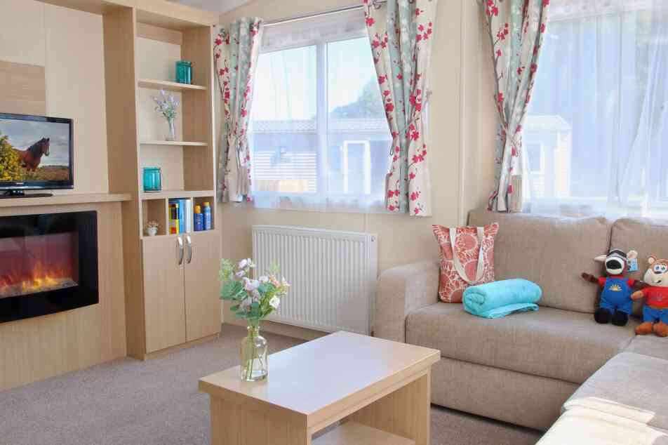 A cozy living room featuring a light-colored couch adorned with decorative pillows, a small coffee table, and a stylish electric fireplace. There is a television on a shelf, surrounded by decorative teal glass vases. Window curtains with floral patterns frame bright windows, letting in natural light. A vase with flowers sits on the coffee table, adding a touch of freshness to the space.