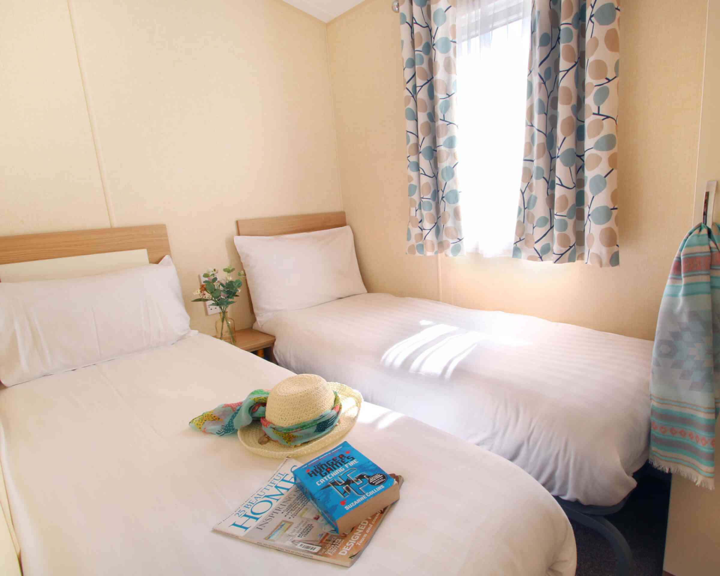 A cozy room with two single beds, light-colored bedding, and a window draped with patterned curtains. A hat and a stack of magazines are placed on one bed, while a towel hangs on the wall nearby. Natural light brightens the space.