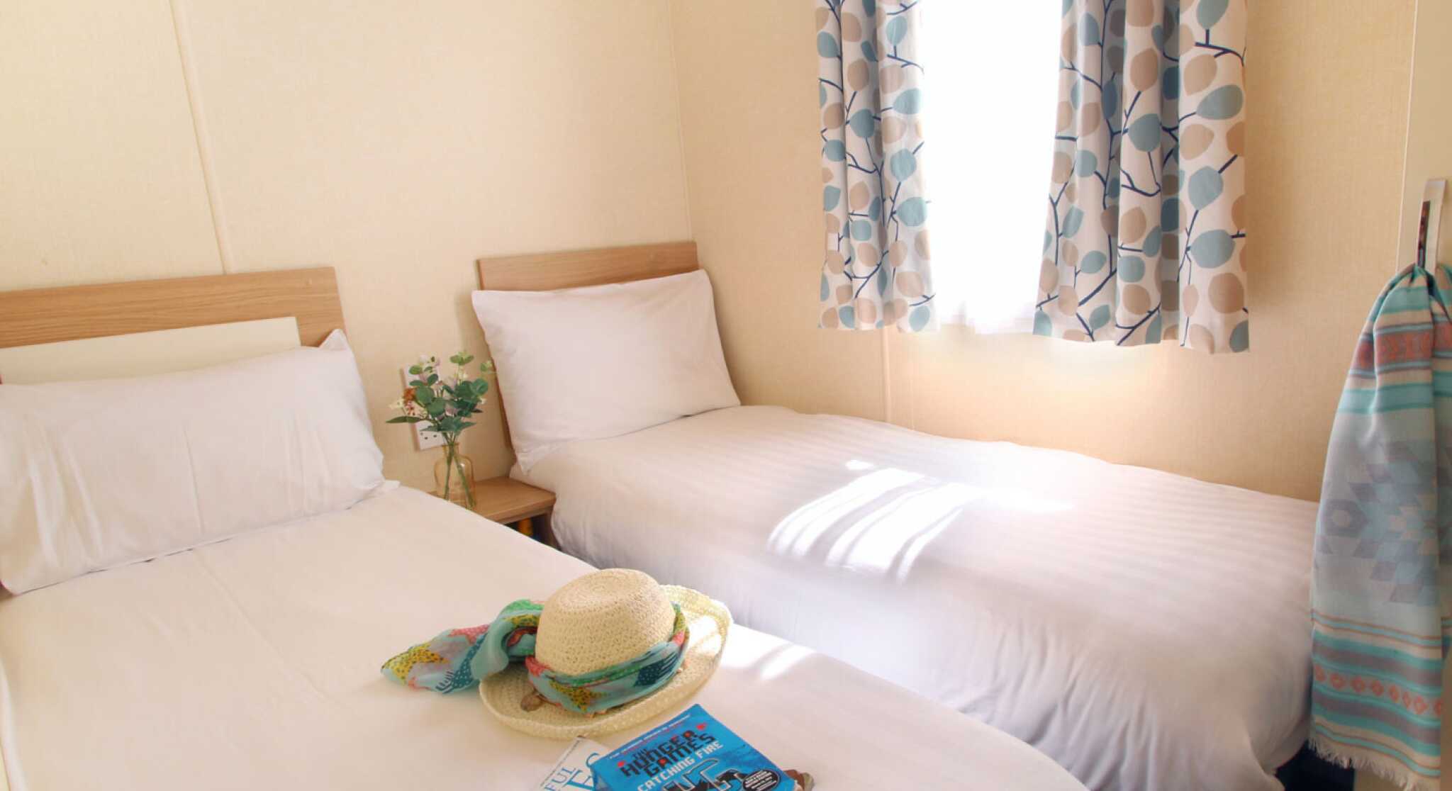 A cozy room with two single beds, light-colored bedding, and a window draped with patterned curtains. A hat and a stack of magazines are placed on one bed, while a towel hangs on the wall nearby. Natural light brightens the space.