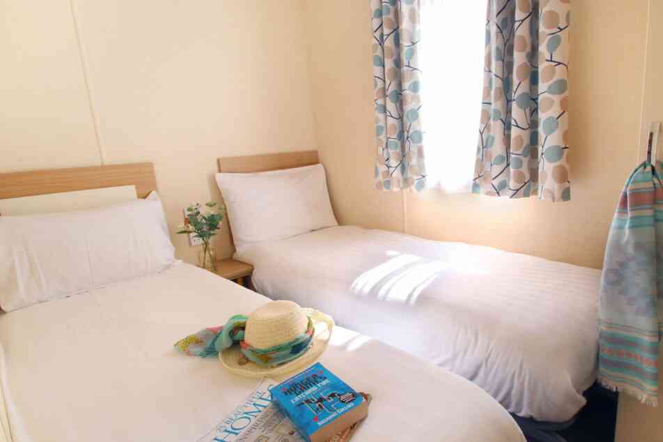A cozy room with two single beds, light-colored bedding, and a window draped with patterned curtains. A hat and a stack of magazines are placed on one bed, while a towel hangs on the wall nearby. Natural light brightens the space.
