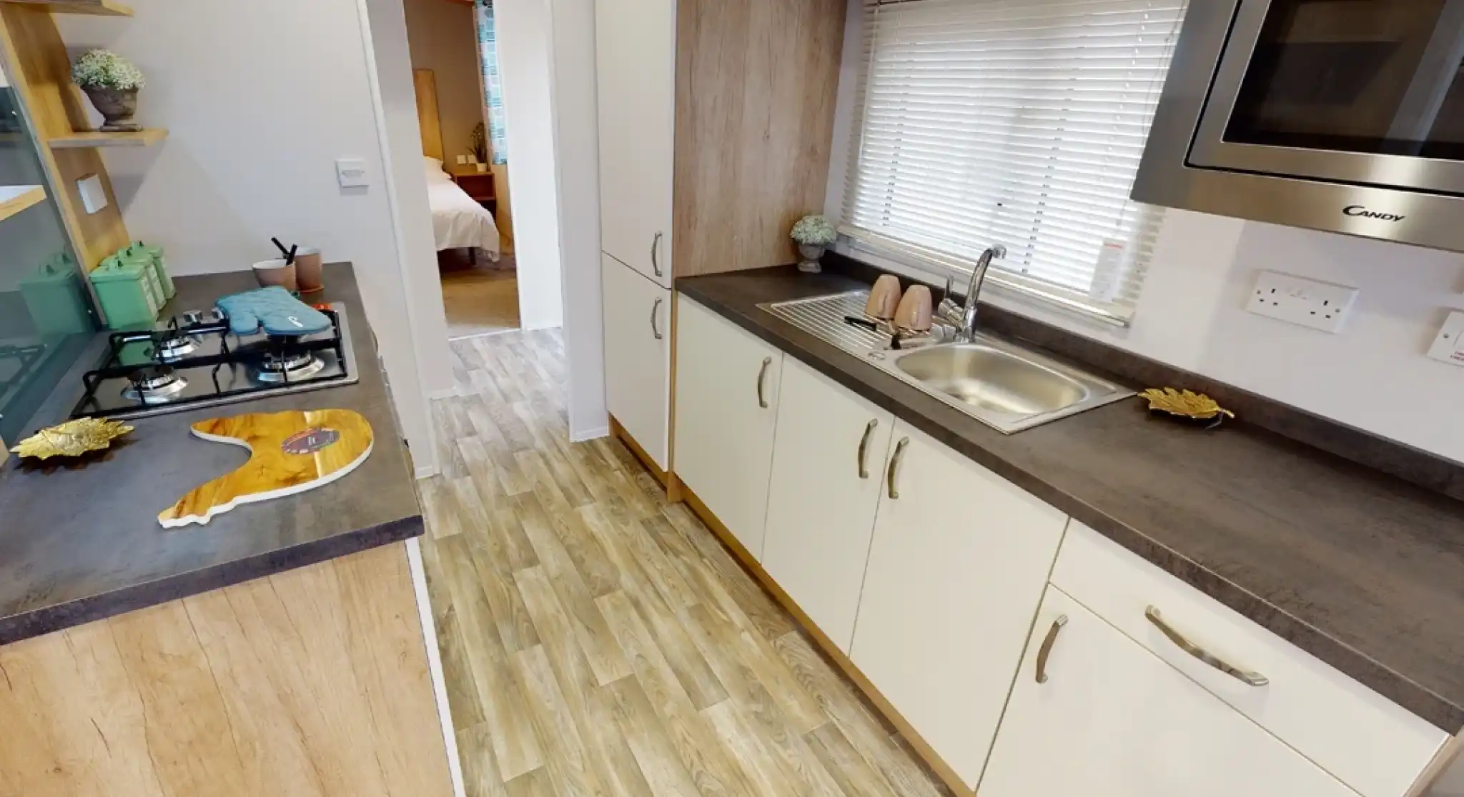 A modern kitchen with light wood cabinetry and a stainless steel sink. The countertop features a gas stove and a microwave overhead. Natural light filters through the window, illuminating the space. In the background, a doorway leads to a bedroom.