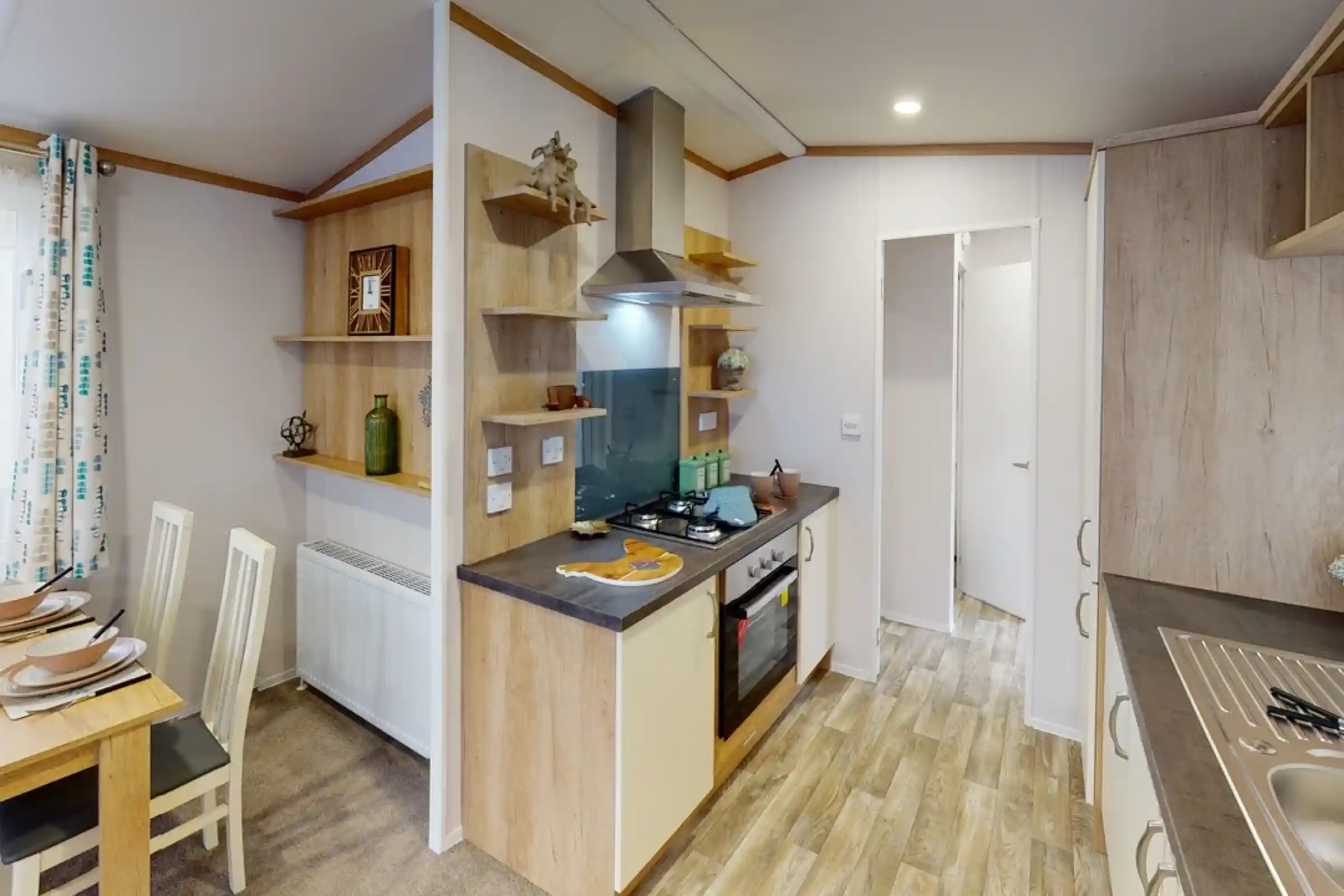 A modern kitchen featuring light wooden cabinetry, a gas stove, and an extractor hood. Open shelves display decorative items and kitchenware. A dining area with a small table and white chairs is visible. The flooring is a light wood laminate.