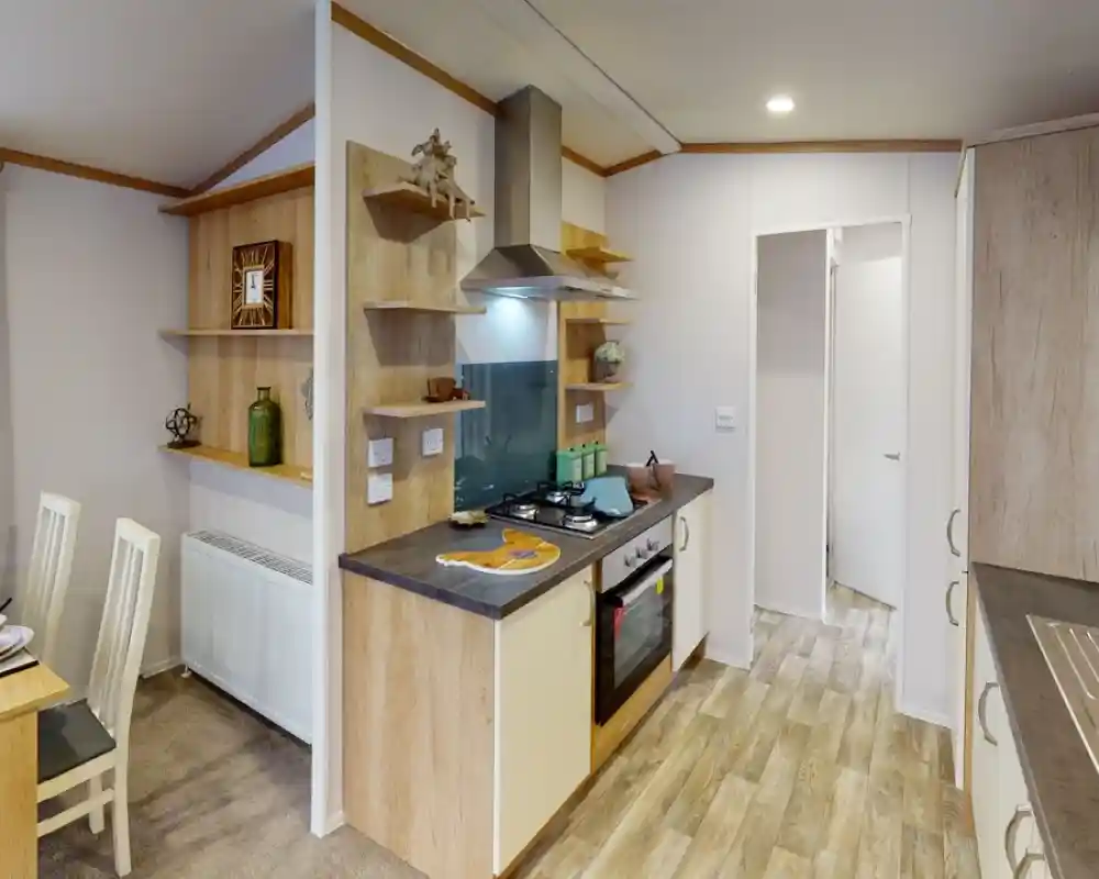 A modern kitchen featuring light wooden cabinetry, a gas stove, and an extractor hood. Open shelves display decorative items and kitchenware. A dining area with a small table and white chairs is visible. The flooring is a light wood laminate.