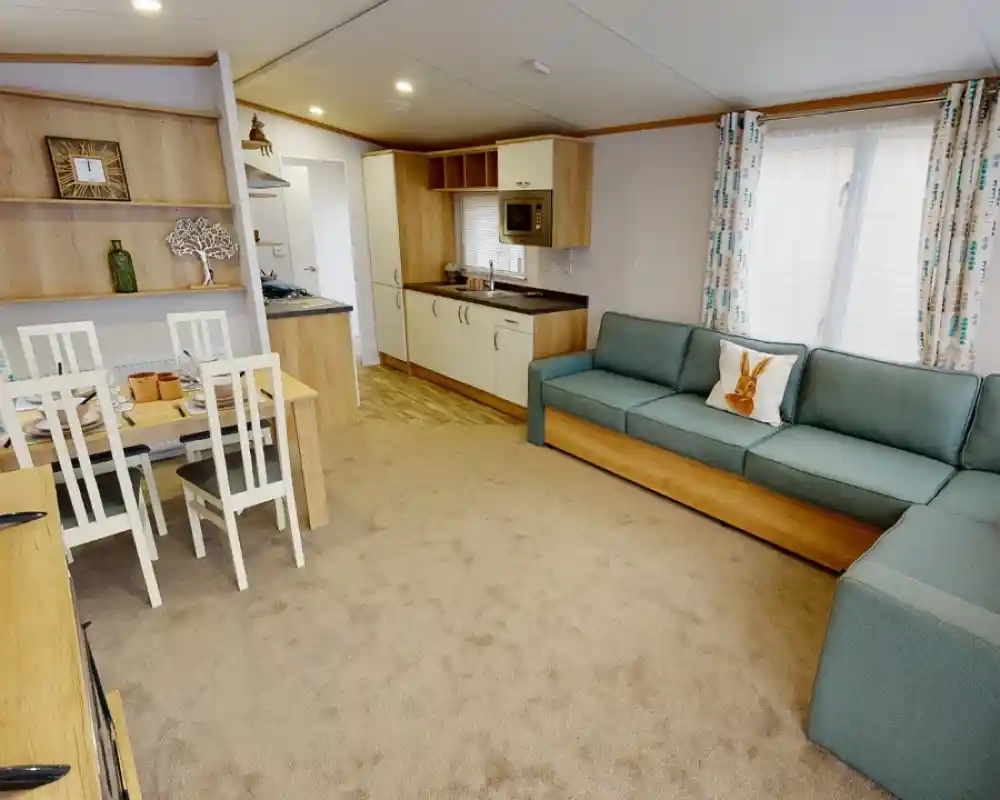 A cozy living space featuring a light-colored carpet, a blue sectional sofa, and a wooden dining table with white chairs. The kitchen area includes modern appliances and wooden cabinetry, illuminated by natural light through a window with patterned curtains.