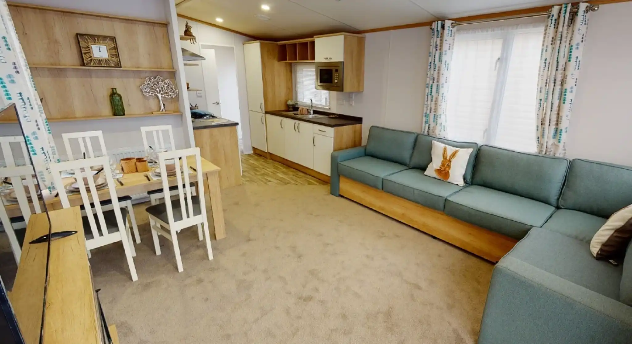 A cozy living space featuring a light-colored carpet, a blue sectional sofa, and a wooden dining table with white chairs. The kitchen area includes modern appliances and wooden cabinetry, illuminated by natural light through a window with patterned curtains.