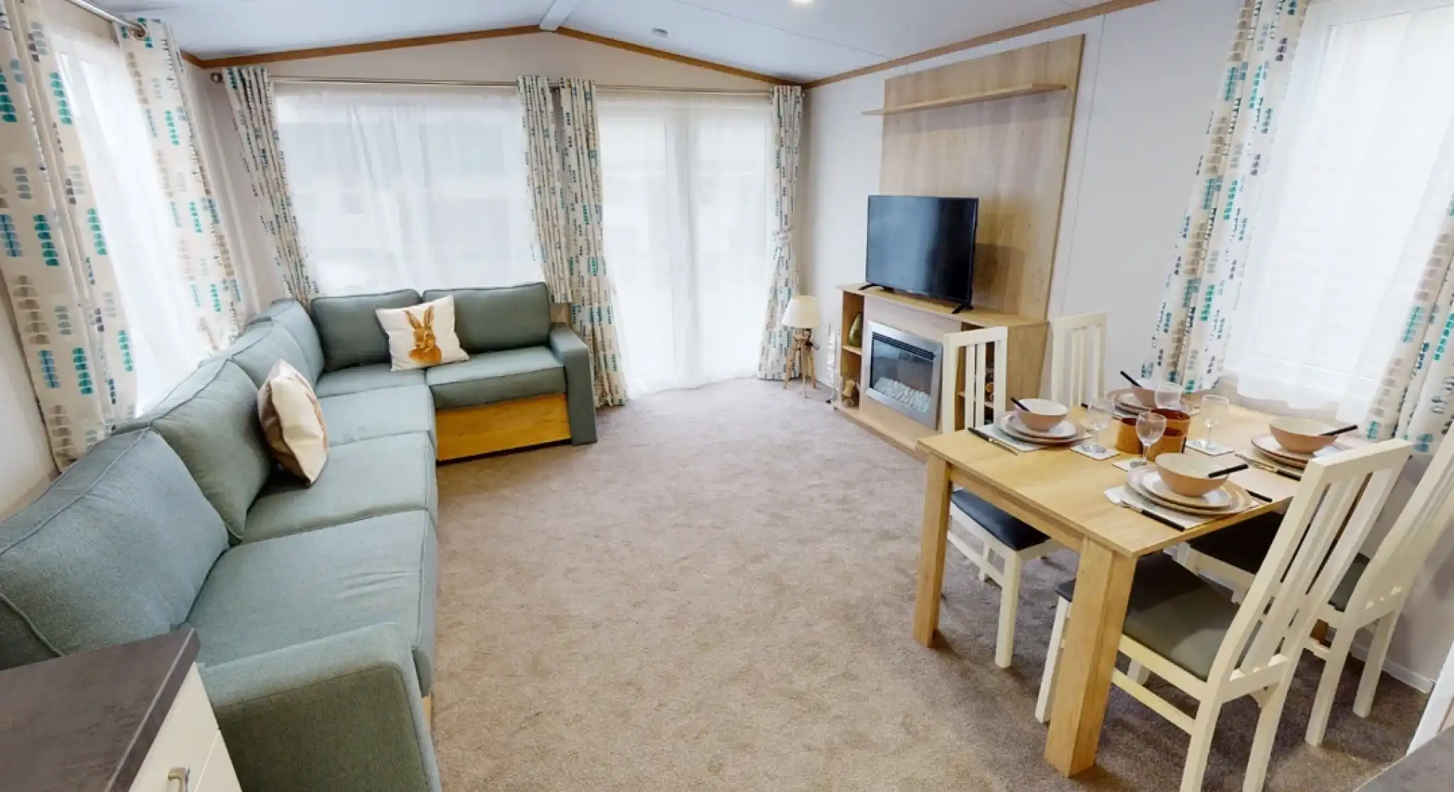 A cozy living area featuring a light blue sectional sofa, a wooden dining table set for four, and a large TV mounted on the wall. Natural light streams in through large windows with patterned curtains. The carpet is beige, creating a warm atmosphere.