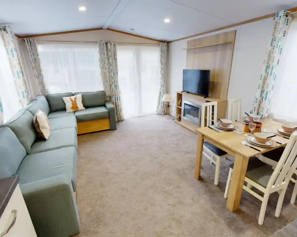 A cozy living area featuring a light blue sectional sofa, a wooden dining table set for four, and a large TV mounted on the wall. Natural light streams in through large windows with patterned curtains. The carpet is beige, creating a warm atmosphere.