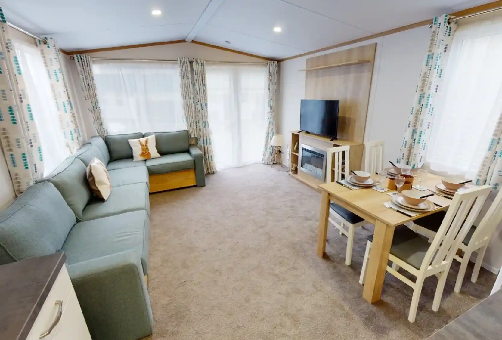 A cozy living area featuring a light blue sectional sofa, a wooden dining table set for four, and a large TV mounted on the wall. Natural light streams in through large windows with patterned curtains. The carpet is beige, creating a warm atmosphere.