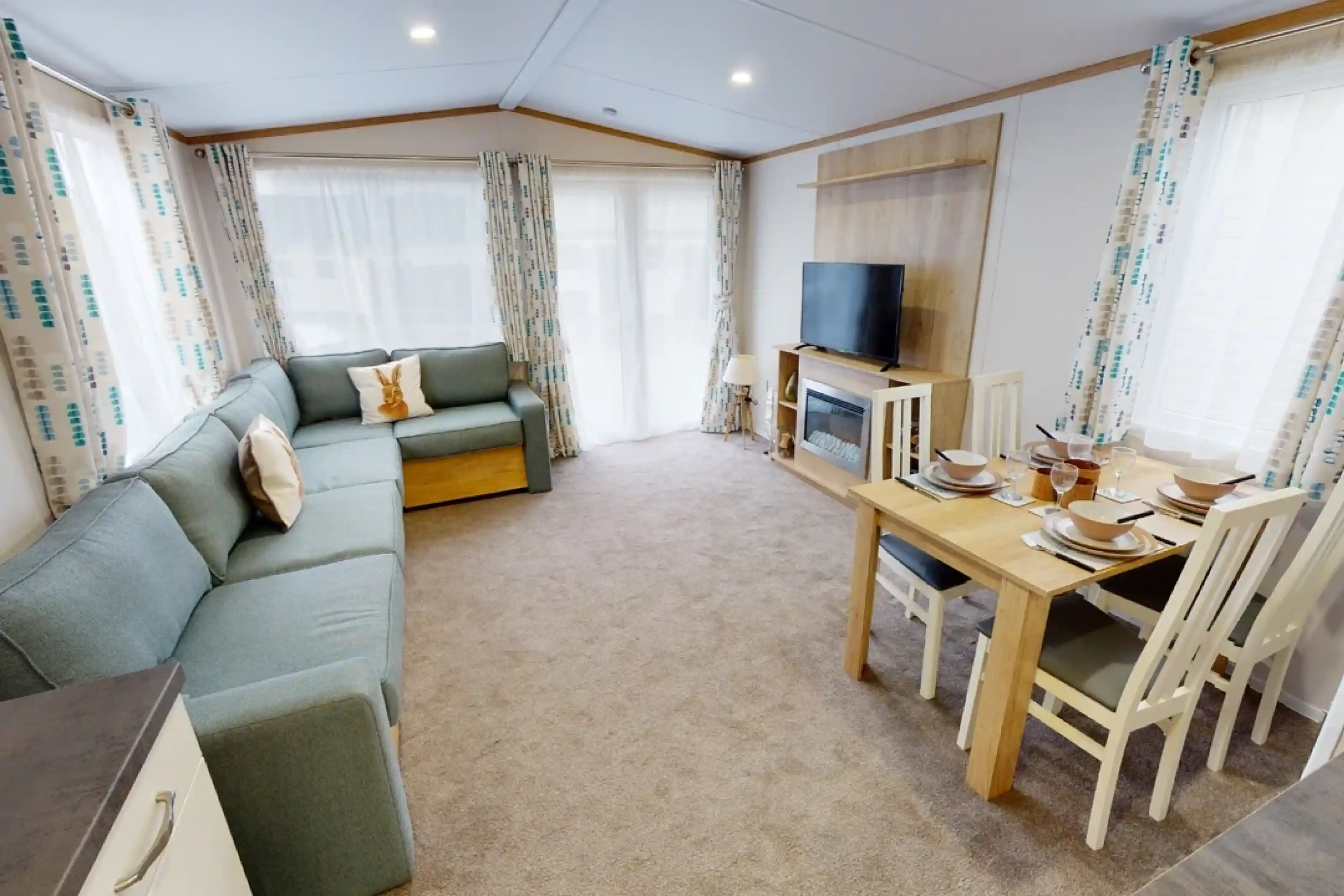 A cozy living area featuring a light blue sectional sofa, a wooden dining table set for four, and a large TV mounted on the wall. Natural light streams in through large windows with patterned curtains. The carpet is beige, creating a warm atmosphere.