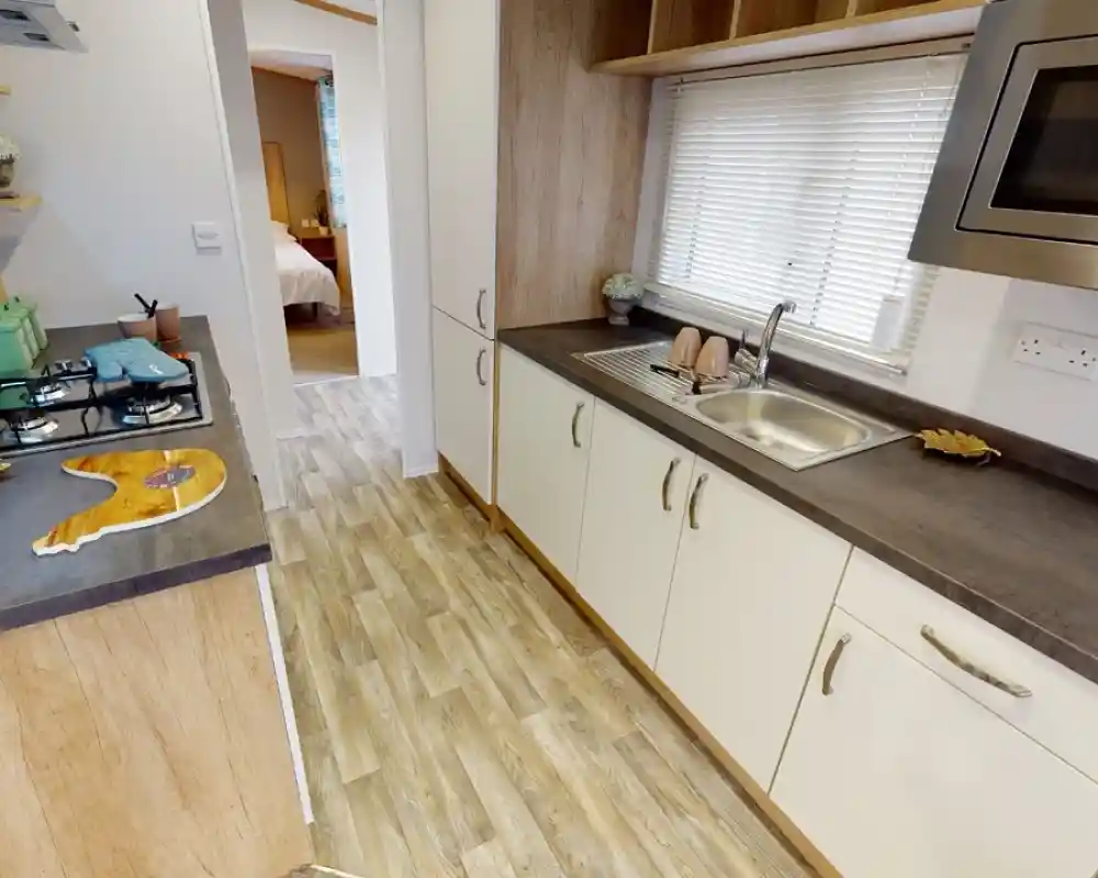A modern kitchen featuring wooden cabinetry, a stainless steel sink, and a gas stove. Natural light streams in through a window with blinds, and decorative items are placed on the shelves. A doorway leads to a bedroom area in the background.