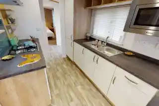 A modern kitchen featuring wooden cabinetry, a stainless steel sink, and a gas stove. Natural light streams in through a window with blinds, and decorative items are placed on the shelves. A doorway leads to a bedroom area in the background.