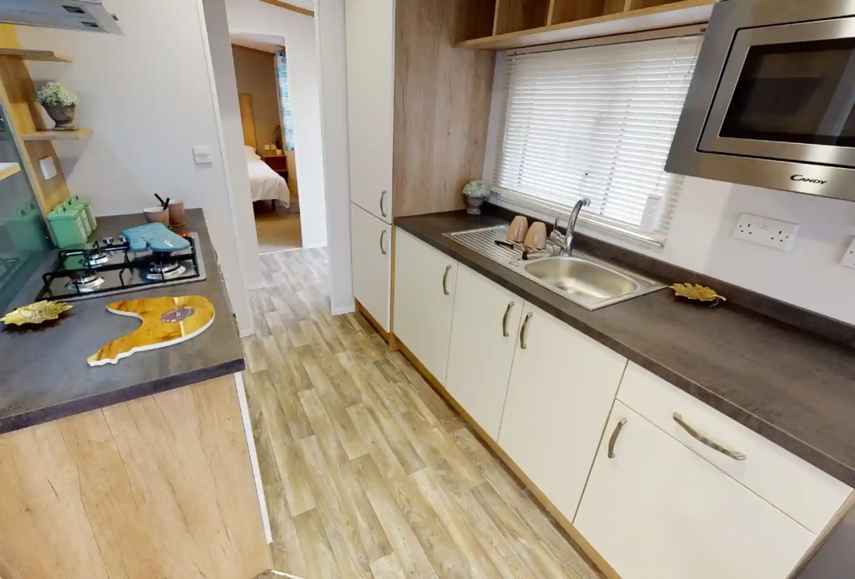 A modern kitchen featuring wooden cabinetry, a stainless steel sink, and a gas stove. Natural light streams in through a window with blinds, and decorative items are placed on the shelves. A doorway leads to a bedroom area in the background.