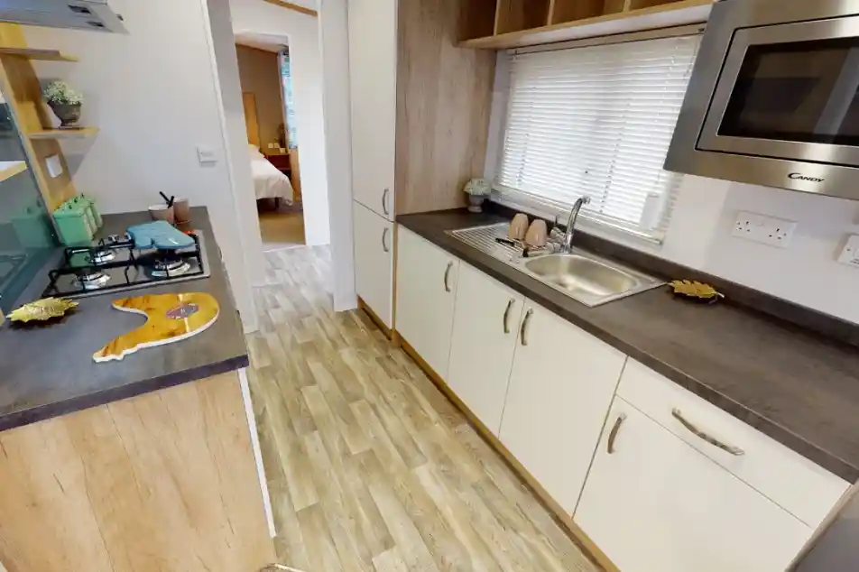 A modern kitchen featuring wooden cabinetry, a stainless steel sink, and a gas stove. Natural light streams in through a window with blinds, and decorative items are placed on the shelves. A doorway leads to a bedroom area in the background.