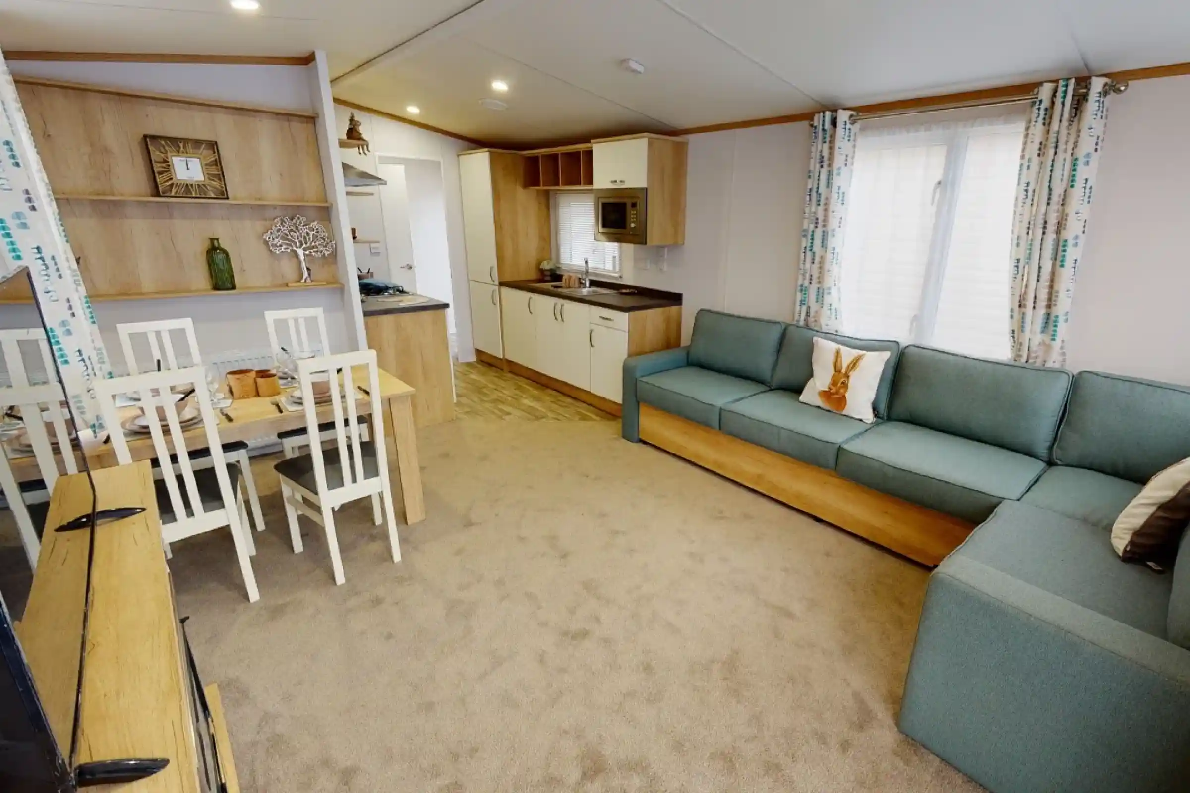 A modern living area featuring a light-colored carpet, a turquoise sectional sofa, a wooden dining table with white chairs, and a compact kitchen with beige cabinetry and an oven. Natural light filters through the windows adorned with patterned curtains.