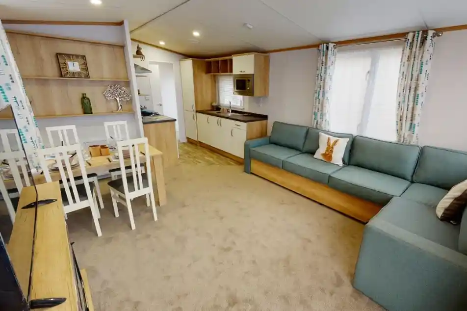 A modern living area featuring a light-colored carpet, a turquoise sectional sofa, a wooden dining table with white chairs, and a compact kitchen with beige cabinetry and an oven. Natural light filters through the windows adorned with patterned curtains.