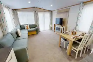 A cozy living space featuring a light blue sectional sofa, a wooden dining table set for four, and a flat-screen TV mounted on the wall. Large windows are adorned with patterned curtains, allowing natural light to fill the room. The flooring is carpeted, creating a warm and inviting atmosphere.