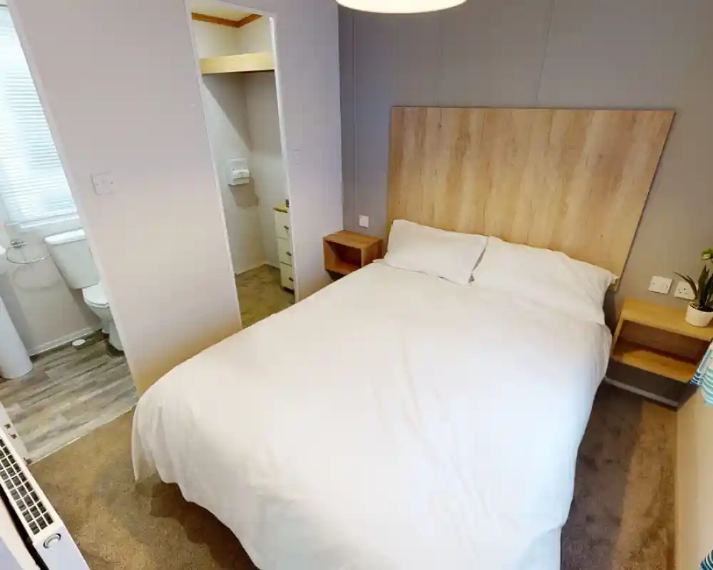 Cozy bedroom with a double bed, wooden headboard, and bedside tables. A small en-suite bathroom is visible in the background. Soft lighting and neutral decor create a welcoming atmosphere.