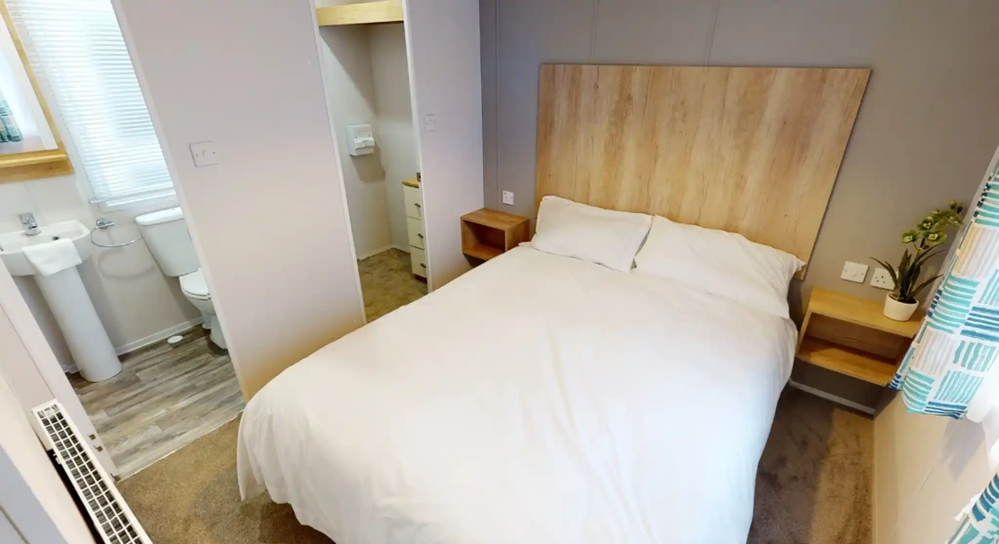 Cozy bedroom with a double bed, wooden headboard, and bedside tables. A small en-suite bathroom is visible in the background. Soft lighting and neutral decor create a welcoming atmosphere.