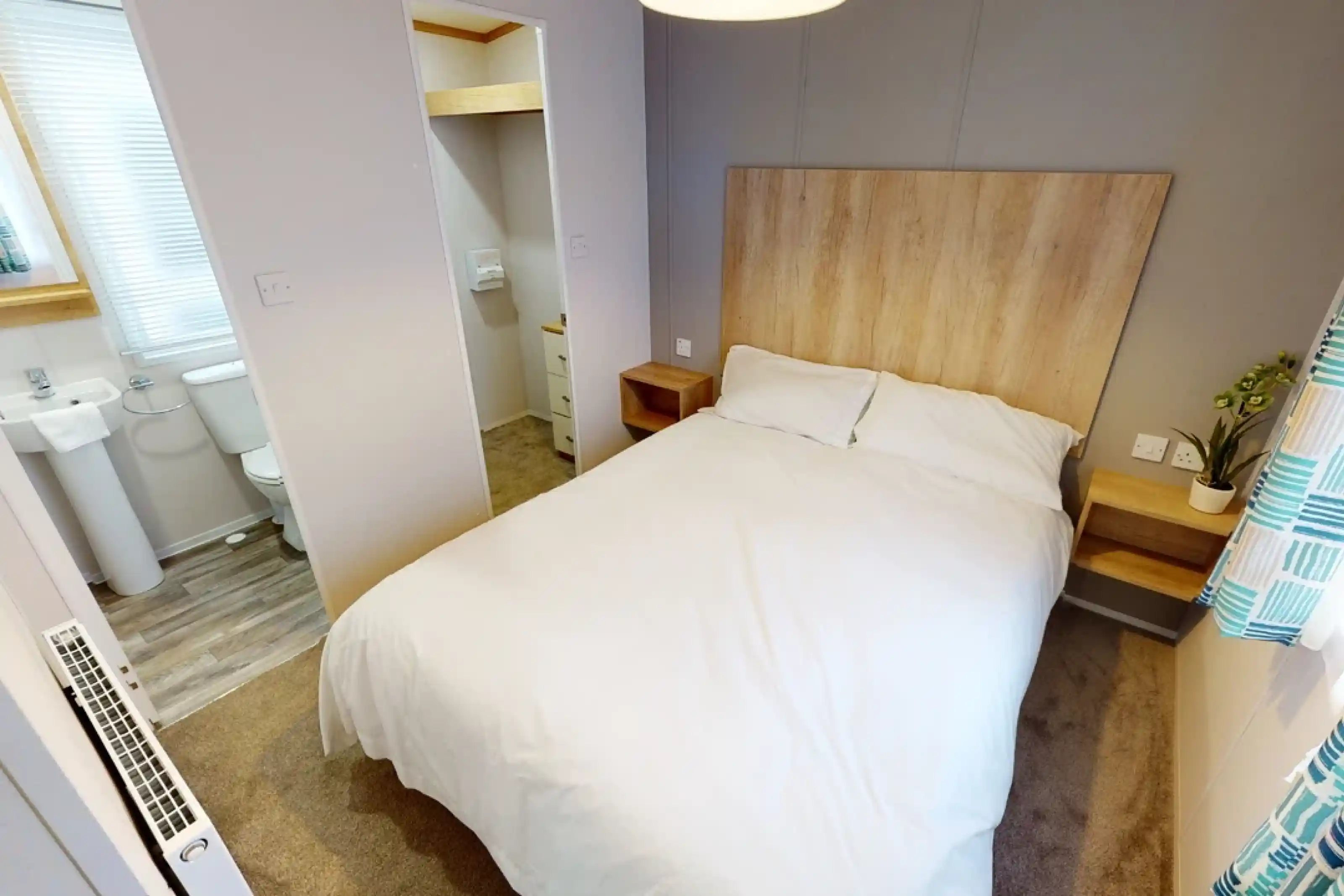 Cozy bedroom with a double bed, wooden headboard, and bedside tables. A small en-suite bathroom is visible in the background. Soft lighting and neutral decor create a welcoming atmosphere.