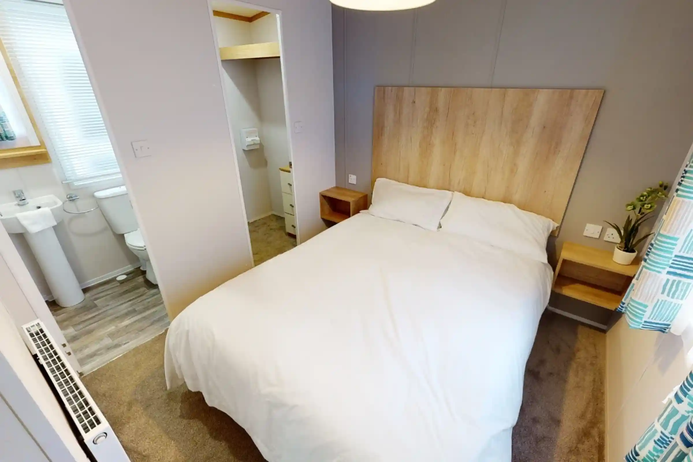 Cozy bedroom with a double bed, wooden headboard, and bedside tables. A small en-suite bathroom is visible in the background. Soft lighting and neutral decor create a welcoming atmosphere.