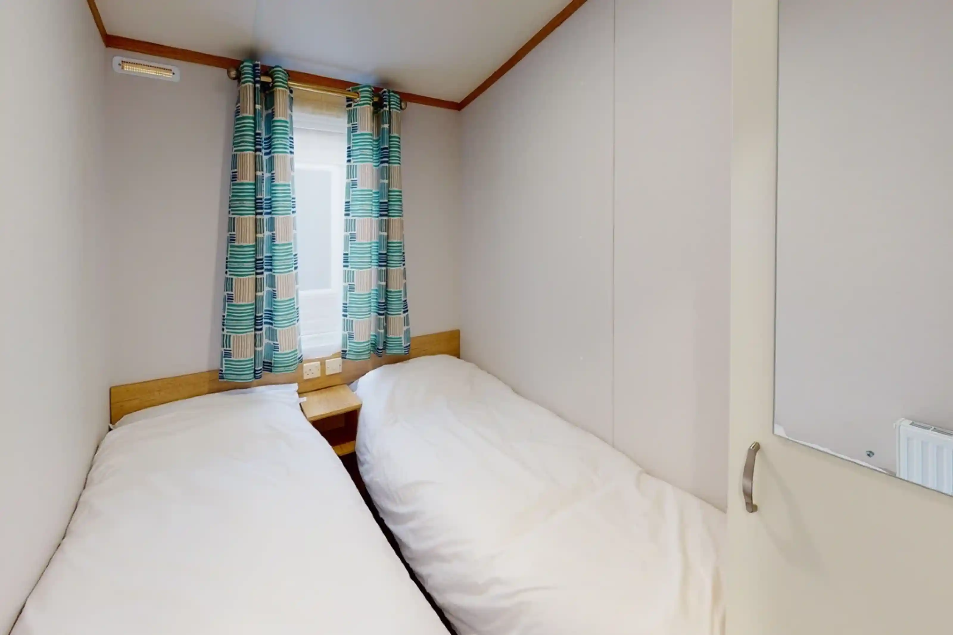 A cozy bedroom featuring two single beds with white bedding. Colorful patterned curtains frame a window, and a small nightstand sits between the beds. The walls are light-colored and have a simple, minimalistic design.