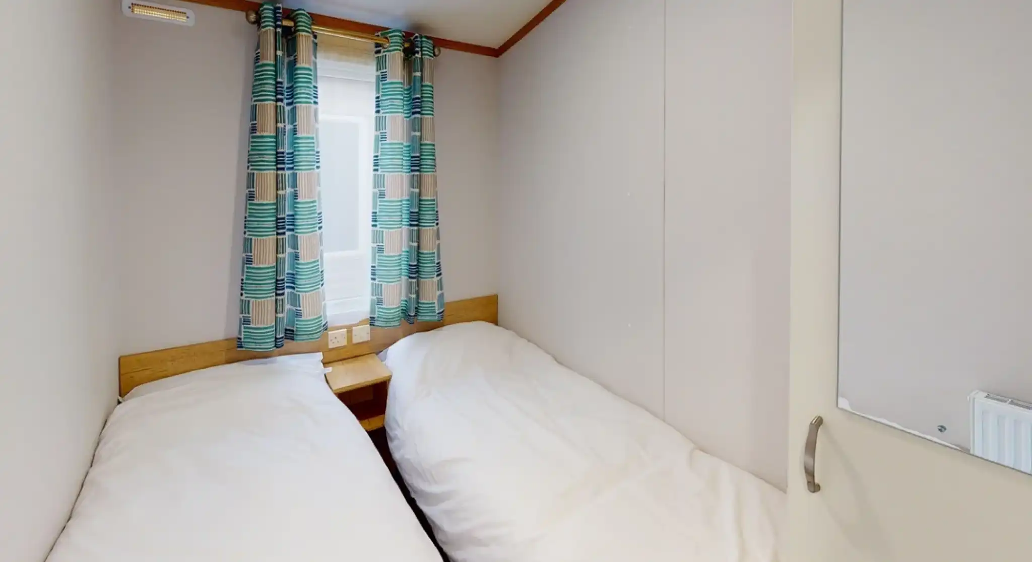 A cozy bedroom featuring two single beds with white bedding. Colorful patterned curtains frame a window, and a small nightstand sits between the beds. The walls are light-colored and have a simple, minimalistic design.