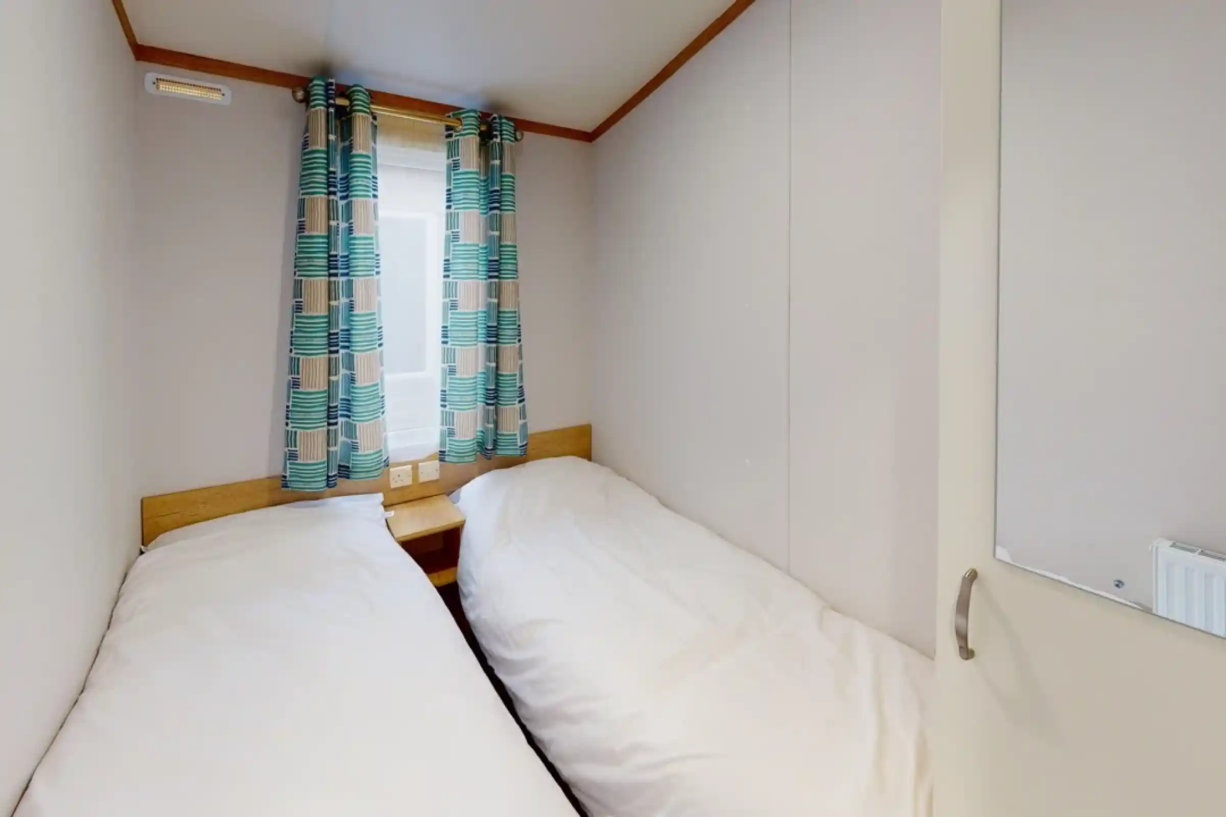 A cozy bedroom featuring two single beds with white bedding. Colorful patterned curtains frame a window, and a small nightstand sits between the beds. The walls are light-colored and have a simple, minimalistic design.