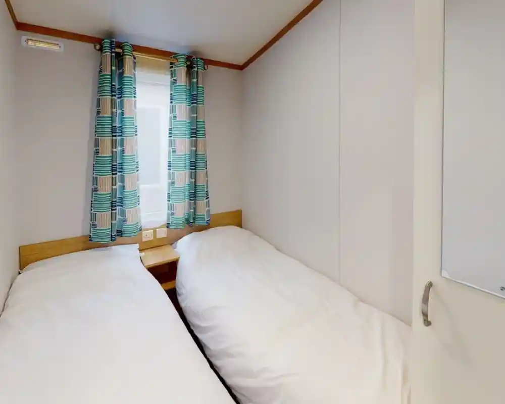 Cozy bedroom with two twin beds positioned side by side, light bedding, and colorful patterned curtains. A window allows natural light, and a mirror is visible on the door.