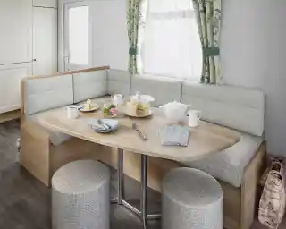 A modern dining area featuring a rounded wooden table with a variety of food, including pastries and a teapot, surrounded by cushioned seating and stools. Natural light enters through a window with floral curtains.