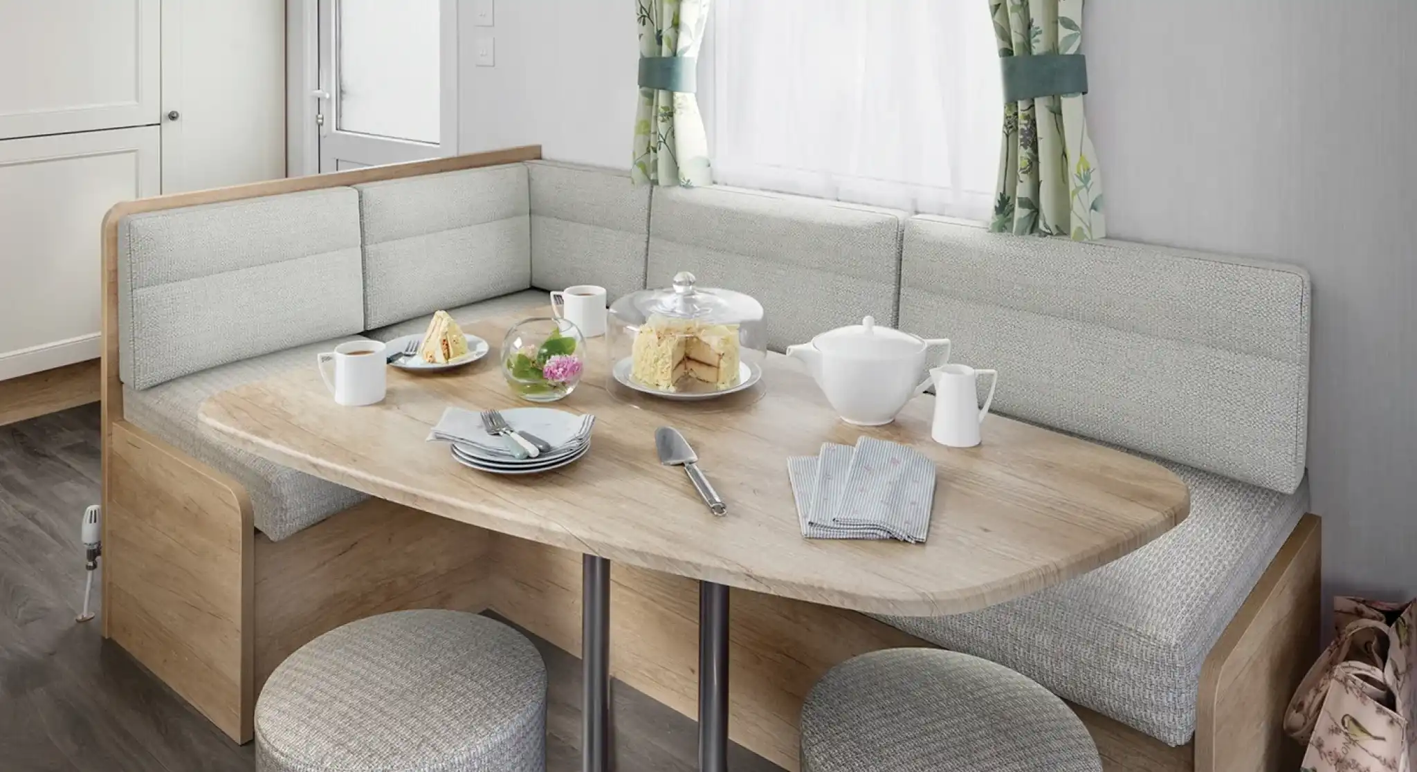 A modern dining area featuring a rounded wooden table with a variety of food, including pastries and a teapot, surrounded by cushioned seating and stools. Natural light enters through a window with floral curtains.