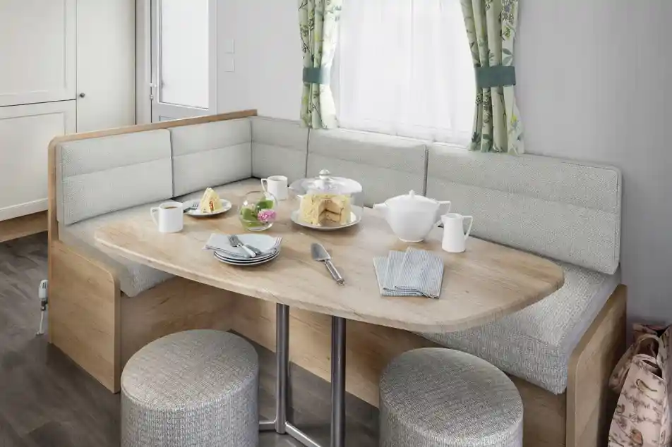 A modern dining area featuring a rounded wooden table with a variety of food, including pastries and a teapot, surrounded by cushioned seating and stools. Natural light enters through a window with floral curtains.