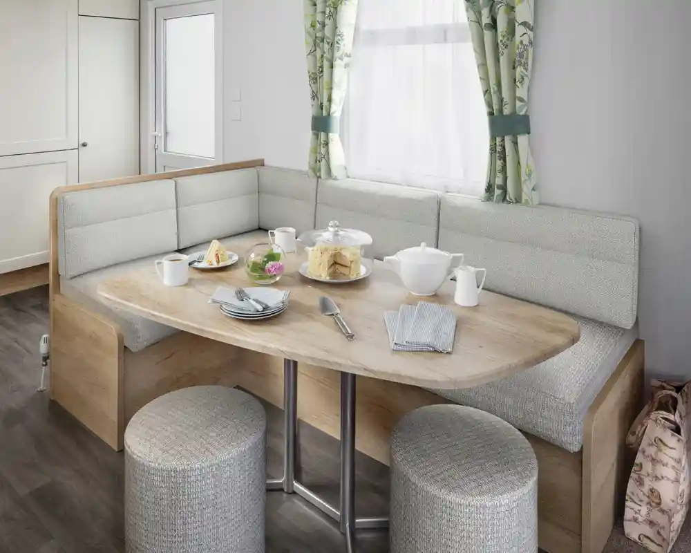 A modern dining area featuring a rounded wooden table with a variety of food, including pastries and a teapot, surrounded by cushioned seating and stools. Natural light enters through a window with floral curtains.