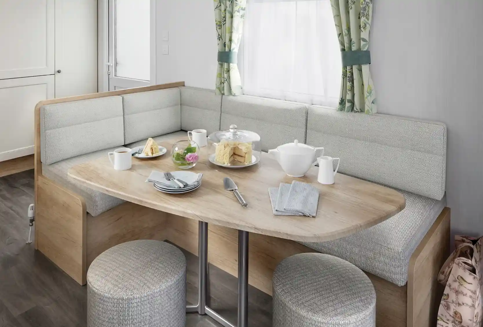 A modern dining area featuring a rounded wooden table with a variety of food, including pastries and a teapot, surrounded by cushioned seating and stools. Natural light enters through a window with floral curtains.