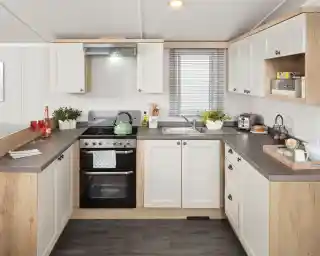 Modern kitchen featuring white cabinets with wooden accents, a stainless steel stove, and a sink. Countertops are dark, and the space includes small plants, dishes, and breakfast items. Natural light filters through a window with blinds.