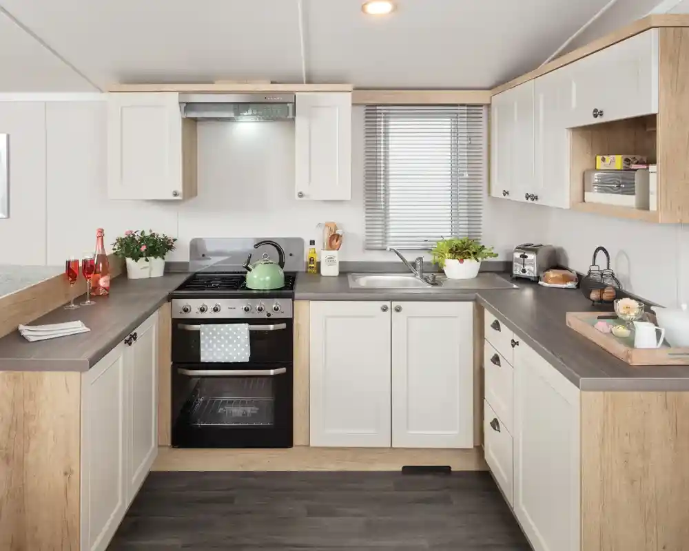 Modern kitchen featuring white cabinets with wooden accents, a stainless steel stove, and a sink. Countertops are dark, and the space includes small plants, dishes, and breakfast items. Natural light filters through a window with blinds.
