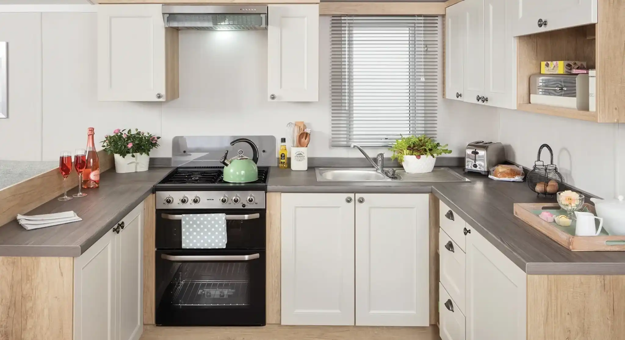 Modern kitchen featuring white cabinets with wooden accents, a stainless steel stove, and a sink. Countertops are dark, and the space includes small plants, dishes, and breakfast items. Natural light filters through a window with blinds.